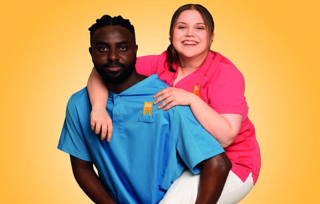 Zwei Auszubildende in SBK-Dienstkleidung lächeln in die Kamera. Einer trägt die andere Huckepack.